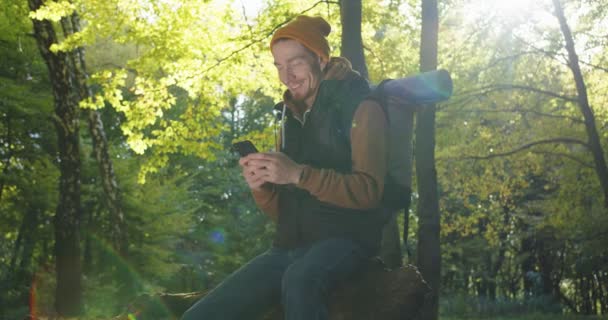 Primer plano de mochilero masculino guapo sentado relajándose en el bosque y mensajes de texto usando smartphone, leyendo mensajes y sonriendo. Concepto de estilo de vida. Hombre excursionista usando el teléfono móvil en los bosques. — Vídeo de stock