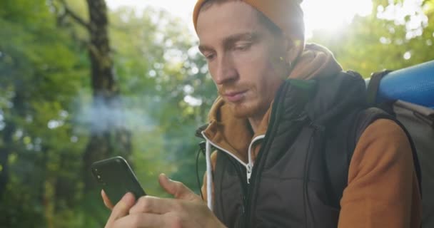 英俊的背包客坐在森林里放松，用智能手机发短信，阅读短信和微笑的形象。旅行生活方式的概念。人类徒步旅行者在树林里用手机. — 图库视频影像