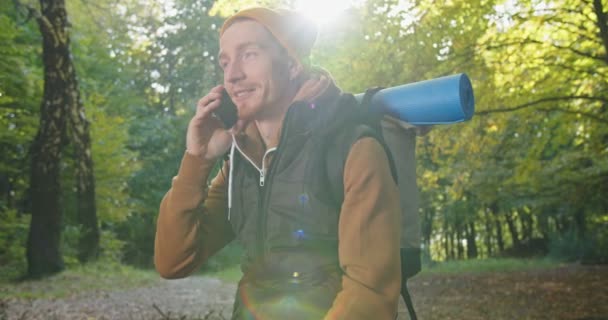 Retrato de mochilero masculino guapo sentado relajándose en el bosque y hablando con un teléfono inteligente, hablando y sonriendo. Concepto de estilo de vida. Hombre excursionista usando el teléfono móvil en los bosques. — Vídeo de stock