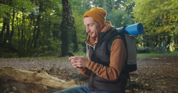 Retrato de mochilero masculino guapo sentado relajándose en el bosque y enviando mensajes de texto usando un teléfono inteligente, leyendo mensajes, charlando, navegando por páginas web y luego riendo. Hombre excursionista usando el teléfono móvil en los bosques. — Vídeo de stock