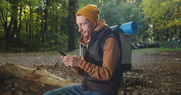 Retrato de mochilero masculino guapo sentado relajándose en el bosque y enviando mensajes de texto usando un teléfono inteligente, leyendo mensajes, charlando, navegando por páginas web y luego riendo. Hombre excursionista usando el teléfono móvil en los bosques. — Vídeo de stock