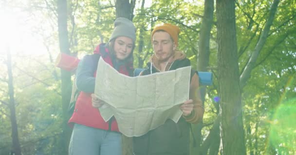 Plano medio de excursionistas interraciales encantadores irritados pareja con mochilas se perdió en el bosque, mirando el mapa de viajes y discutiendo sobre la ruta correcta trek durante el senderismo en la naturaleza de otoño. — Vídeo de stock