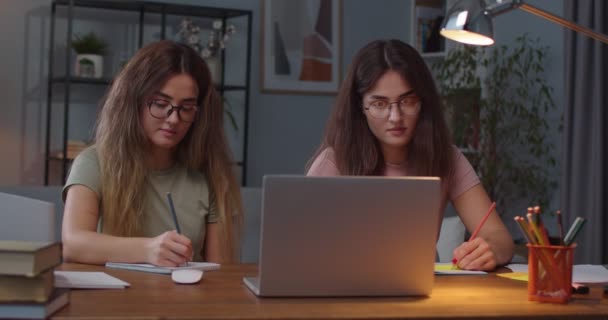 Portret pięknej młodej, szczęśliwej, białej kobiety bliźniaczki siedzącej przy biurku w pokoju, uczącej się z laptopa. Dwie siostry piszące w notatnikach z komputera o edukacji na odległość. Koncepcja e-learningu — Wideo stockowe