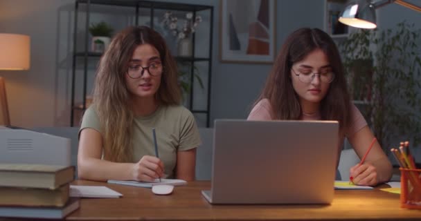 Retrato de dois jovens gêmeos femininos caucasianos felizes sentados à mesa no quarto enquanto escrevem e estudam no laptop. Duas irmãs fazendo anotações em cadernos de informática sobre educação a distância E-learning — Vídeo de Stock