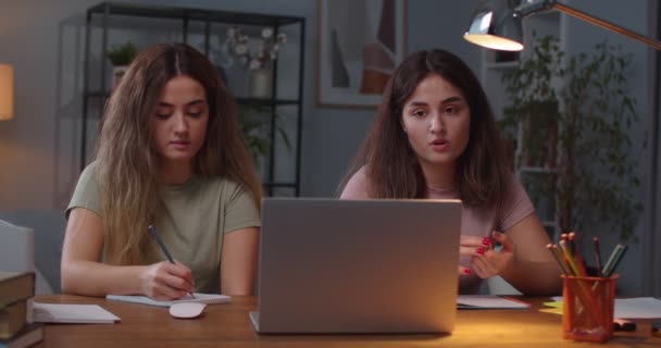 Portret dwóch sióstr bliźniaczek rasy kaukaskiej siedzących w pokoju i uczących się podczas lekcji online na laptopie Ładna kobieta pisząca w notatniku, podczas gdy siostra rozmawiająca na wideo o komputerowej edukacji na odległość — Wideo stockowe