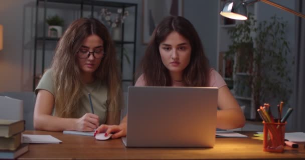 Joven feliz caucásico dos gemelas sentadas en el escritorio en la habitación y estudiando desde el ordenador portátil. Hermanas gemelas aprendiendo en casa. Mujer escribiendo en la computadora mientras su hermana escribe en un libro de copybook. Concepto de estudio — Vídeo de stock