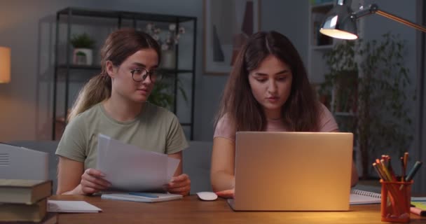 白种人的孪生姐妹坐在舒适的房间里靠笔记本电脑和打字学习。女性在电脑上浏览，并与持有证件的孪生姐妹交谈。远程学习概念 — 图库视频影像