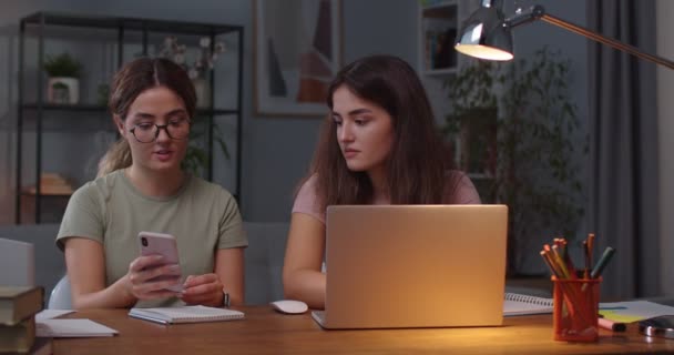 Portrét bělošských dvojčat sedících v pokoji a mluvících při používání přístrojů. Žena píše na notebook, zatímco její dvojče hledá internet na smartphonu a něco jí ukazuje. Koncept volného času — Stock video