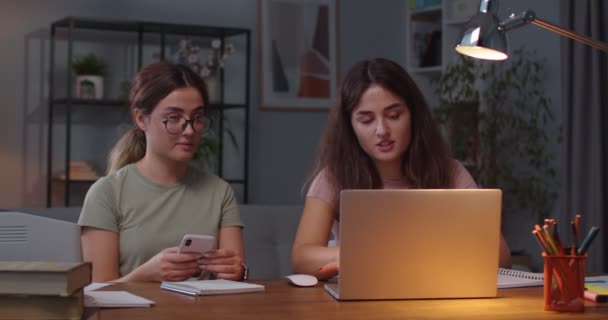 在舒适的房间里，白种人孪生姐妹坐在办公桌前，带着笔记本电脑和智能手机的肖像。女人们在电脑上打耳光，并与正在用手机发短信的孪生姐妹交谈。室内概念 — 图库视频影像