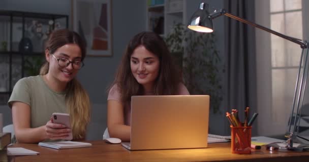 Счастливые кавказские близнецы сидят в комнате и разговаривают, используя устройства. Красивая женщина просматривает ноутбук, в то время как ее сестра ищет в интернете на смартфоне и показывает ей что-то. Концепция отдыха — стоковое видео