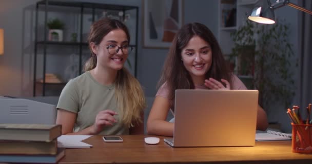 Białe, piękne, młode bliźniaczki machające dłońmi i uśmiechnięte podczas rozmowy przez Internet na laptopie. Portret wesołych sióstr odbywających wideo czat na komputerze w przytulnym domu. Koncepcja czasu wolnego — Wideo stockowe