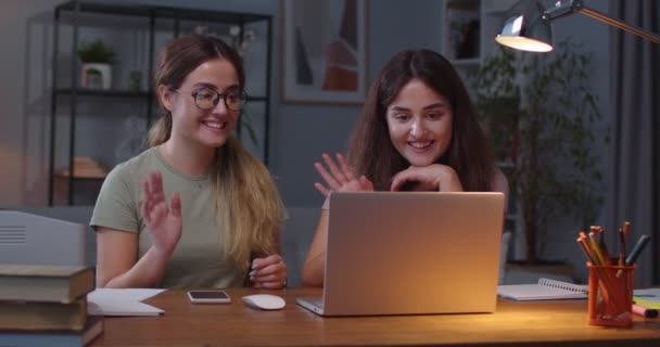Ritratto di belle giovani gemelle caucasiche felici che salutano le mani e sorridono mentre parlano in videochiamata sul computer portatile. Gioioso sorelle video chat sul computer a casa accogliente. Concetto di tempo libero — Video Stock