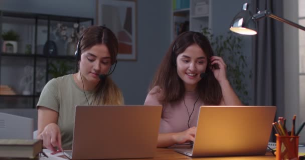 Radosne siostry bliźniaczki rasy kaukaskiej siedzące w zagłówkach przy stole w pokoju i mówiące na wideokonferencji na laptopach Kobiety biorące lekcje online podczas pracy na komputerach w domu Praca na odległość E-learning — Wideo stockowe