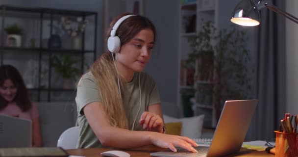 Retrato de feliz jovem caucasiana batendo no laptop e ouvir música em fones de ouvido, enquanto sua irmã gêmea sentado no sofá no fundo e navegando no smartphone em casa. Conceito de lazer — Vídeo de Stock