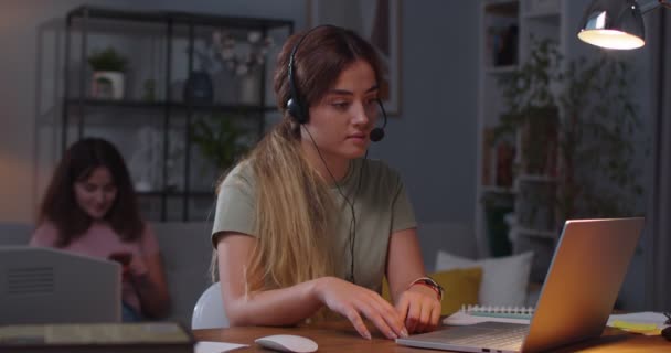 Retrato de la joven mujer caucásica en auriculares hablando en video llamada en línea en el ordenador portátil, mientras que su hermana gemela sentado mensajes de texto en el teléfono inteligente en el fondo en casa. Concepto de educación a distancia — Vídeo de stock