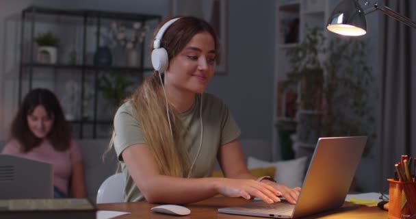 Χαρούμενη Καυκάσια γυναίκα περιήγηση στο laptop, ενώ ακούτε μουσική με ακουστικά και αστεία κίνηση και γυρίζοντας πίσω στη δίδυμη αδελφή της στο παρασκήνιο που χτυπάει στο smartphone. Αρχική ιδέα — Αρχείο Βίντεο