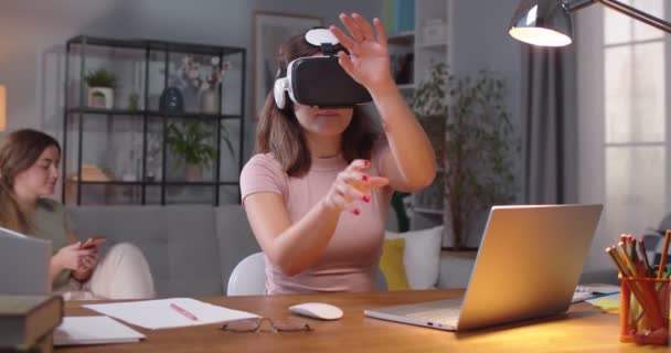 Белая красивая молодая женщина в очках VR с использованием футуристических технологий и жесты в воздухе в то время как сестра-близнец на заднем плане использует смартфон Портрет женщины в 3D виртуальной реальности гарнитура — стоковое видео