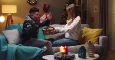 Kanepede oturan mutlu karı-kocanın orta planı. Kapalı gözlü erkek arkadaş hediyesini bekliyor. Kız arkadaş büyük güzel bir Noel hediyesi veriyor ve sarılıyor. Aile tatili kavramı.