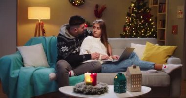 Karı kocanın koltukta oturmasının orta planı. Sevgilisi kız arkadaşına yardım ediyor. Tatil kutlamalarını bilgisayarda yazıyor ve gülümsüyor. Aile kavramı, mükemmel ilişkiler, Noel zamanı..