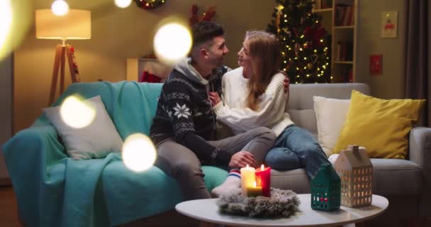 Piano medio di coppia fidanzato e fidanzata seduti sul divano, abbracci, baci, guardarsi l'un l'altro, sorridente, umore festivo in soggiorno. Concetto di vacanze in famiglia, relazioni, Natale. — Video Stock