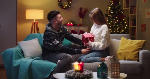 Środkowy plan szczęśliwej pary mąż i żona siedzą na kanapie, przytulają się. Prezent na otwarcie dziewczyny, czerwone pudełko z prezentami. Pojęcie wakacji rodzinnych, idealne związki w okresie Bożego Narodzenia. — Wideo stockowe
