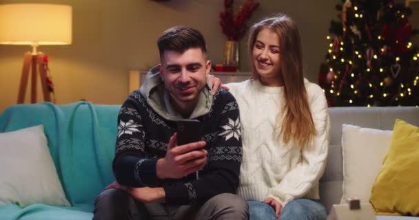 Primer plano de la feliz pareja marido y mujer sentado en el sofá, llamando a la webcam, vídeo chat con amigos utilizando el teléfono, saludando de la mano, sonriendo en la víspera de Navidad en el estado de ánimo festivo en la sala de estar. — Vídeo de stock