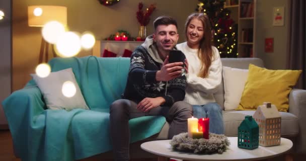 Средний план счастливой пары муж и жена сидят на диване, звонят на веб-камеру, общаются с друзьями по телефону, машут рукой, улыбаются в канун Рождества в праздничном настроении в гостиной. — стоковое видео