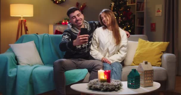Plano medio de la feliz pareja marido y mujer sentado en el sofá, llamando a la webcam, vídeo chat con amigos utilizando el teléfono, besos, abrazos, sonriendo en la víspera de Navidad en el estado de ánimo festivo en la sala de estar. — Vídeo de stock