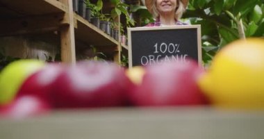 Şapkalı, şapkalı, meyveli seranın arkasında duran, elinde% 100 organik ahşap tabela tutan ve kameraya gülümseyen çekici olgun bir kadın. Hobi tarım kavramı.