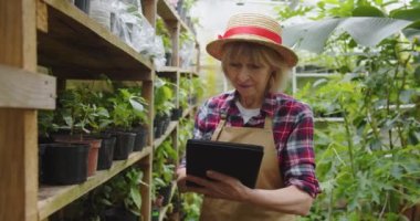 Şapkalı, serada yürüyen, elinde tabletle bitkilerin denetimini yapan çekici, olgun bir kadın. Seradaki güzel pozitif kadın biyologun portresi. Hobi tarım kavramı.