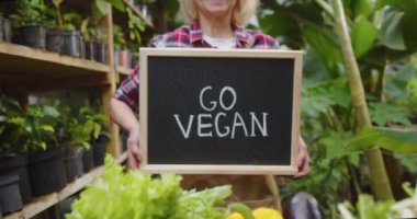 Yetişkin kadınların ellerinde ahşap tabela olan ellerini yakın çekimde vejetaryen ol, seranın arkasındaki kutuda sebzelerle birlikte kameraya göster. Serada poz veren güzel bir kadın biyolog. Hobi tarım kavramı.