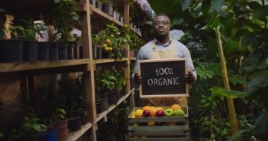 Gözlüklü Afrikalı Amerikalı bir adamın kutunun arkasında elinde meyvelerle dikilip, üzerinde organik tahta tabela tutarak kameraya gösterdiği bir fotoğraf. Çiçekçilik, işgal ve iş konsepti.