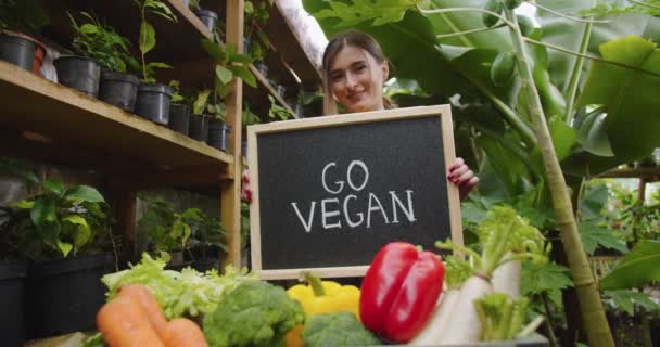 Primo piano di affascinante donna bionda in piedi in serra dietro la scatola con verdure, tenendo segno di legno andare vegan a bordo, mostrando alla fotocamera e sorridente. Hobby concetto di agricoltura. — Video Stock