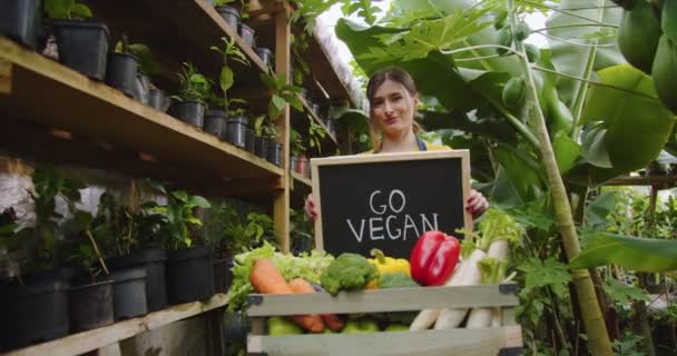 Middle shot of charming blonde woman standing in greenhouse behind box with vegetables, holding wooden sign go vegan on board, showing to camera and smiling. Hobby farming concept. — Stock Video
