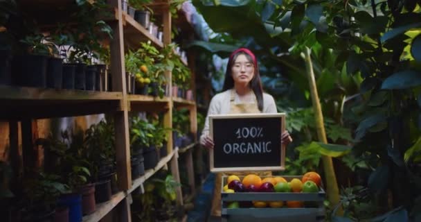 Inquadratura centrale di donna asiatica in occhiali in piedi in serra dietro scatola con frutta, tenendo segno di legno organico, mostrando alla fotocamera e sorridente. Hobby agricoltura, concetto di agricoltura. Telecamera si muove — Video Stock