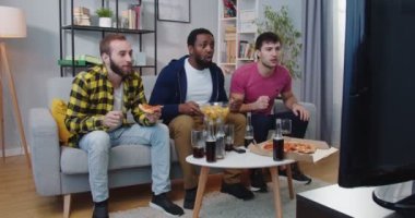 Neşeli, mutlu, beyaz ve Afro-Amerikalı erkekler favori takım maçı için tezahürat yapıyorlar. Erkek arkadaşlar spor kanalında abur cubur ve içeceklerle televizyon izliyorlar. Eğlence konsepti. Arkadaşlık.