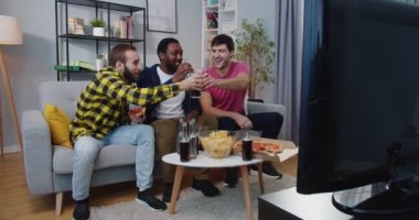 Beyaz ve Afro-Amerikalı genç erkekler oturma odasında kanepede oturup, bardaklarını tokuşturup, TV 'de futbol maçı izliyorlar. En iyi arkadaşlar televizyon karşısında spor kanalıyla konuşurken eğleniyorlar.