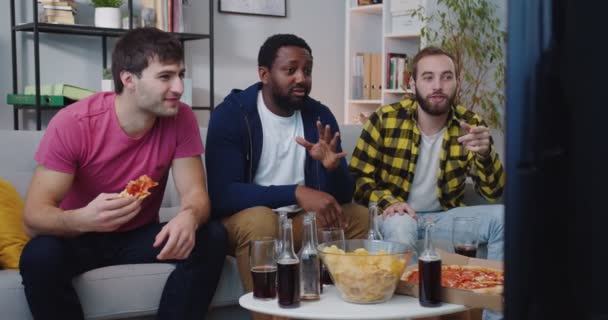 Joyeux jeunes afro-américains et caucasiens amis masculins s'amuser ensemble à la maison assis sur le canapé regarder la chaîne de sport à la télévision avec de la nourriture et des boissons bavarder soutenir l'équipe préférée. Concept de ventilateur — Video