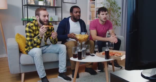 Tres jóvenes felices emocionales carreras mixtas masculinos mejores amigos se reunieron en casa para ver el canal de deportes en la televisión comiendo bocadillos, hablando, apoyando al equipo de fútbol favorito en el concepto de campeonato Juego — Vídeos de Stock