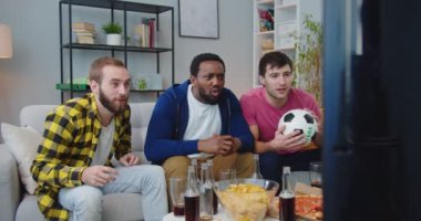 Neşeli Afro-Amerikan ve Kafkasyalı erkekler futbol fanatikleri ile kanepede televizyon seyrederek arkadaş oluyorlar. Genç erkekler çığlık atıyor, sarılıyor ve gol ya da zaferi kutluyor. spor ve eğlence.