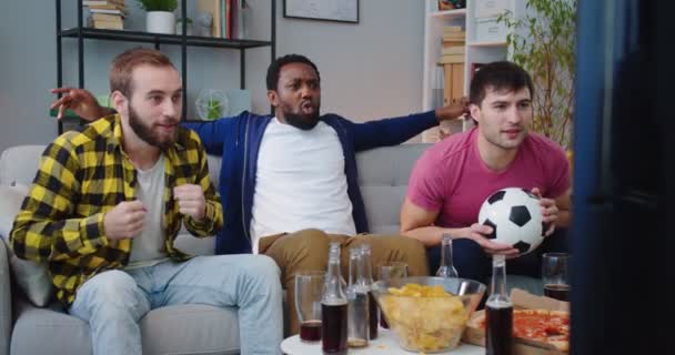 Three handsome happy African American and Caucasian friendly men eating chips snacks and cheering up for football game on TV while sitting on sofa Male friends watching sport channel gathered together — Stock Video