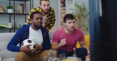 Mutlu Afro-Amerikan ve Kafkas erkek arkadaşları futbol maçı izlerken evdeki kanepede oturmuş, bağırıyor ve gol atmayı kutluyorlar. Erkekler top tutarken spor kanalı izler
