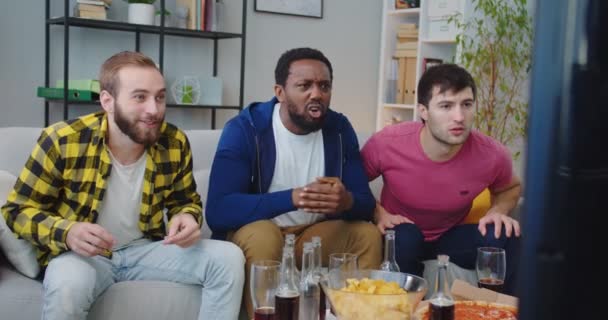 Jóvenes felices afectivos afroamericanos y caucásicos amigos juntos viendo el partido en la televisión en casa. Hombres gritando, abrazando y celebrando la victoria animando al equipo fav. aficionados al fútbol, gol de puntuación — Vídeos de Stock