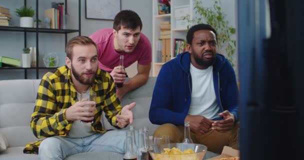 Hombres viendo partidos de fútbol en la tele. Los mejores amigos masculinos ven el partido de fútbol en el canal deportivo en la televisión se reunieron en casa bebiendo y comiendo bocadillos animando al equipo favorito. Concepto de ventilador — Vídeos de Stock