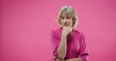 Stüdyoda pembe arka plan üzerinde duran ve bir şeyler düşünen üzgün, tatminsiz yüz ifadesi olan güzel emekli, Kafkasyalı bir kadının portresi. Poz modeli