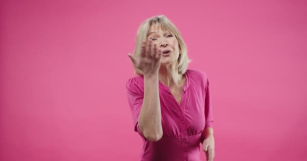 Portré gyönyörű öreg fehér boldog nő így levegő csók, integető kéz, hogy gesztusok örömteli kifejezés pozitív hangulatban áll a stúdióban rózsaszín háttér és mosolygós — Stock videók
