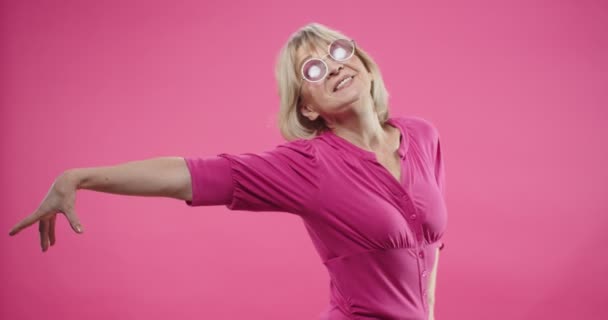Portret wesoły starszy emerytowany biały ładna kobieta w różowych okularach stojący na różowym tle w studio pozowanie i co gesty śmieszne poruszając się w dobrym nastroju i uśmiechając się ze szczęśliwą twarzą — Wideo stockowe