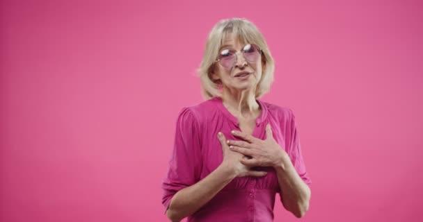 Retrato de idosos muito idosos caucasiano bela mulher com cabelo loiro em rosa óculos elegantes de pé isolado em fundo rosado no estúdio olhando para a câmera, falando fazendo gestos — Vídeo de Stock