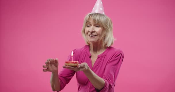Portret van mooie blanke senior vrolijke vrouw in verjaardagshoed houden donut in de handen, blazen kaars op cupcake, geïsoleerd op roze achtergrond Close up portret van oude glimlachende vrouw vieren — Stockvideo