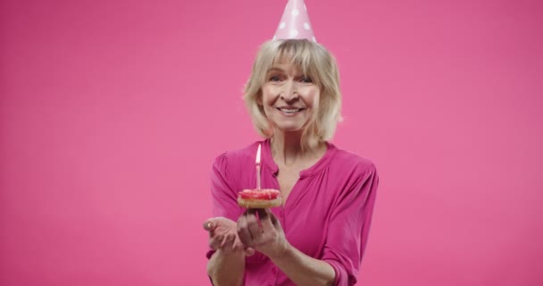 Красива кавказька літня жінка з радісним днем народження носить капелюх, тримаючи пончик у руках, що дмухає свічкою на торті, ізольованому на рожевому рожевому тлі. Закріпіть портрет старої жінки, що святкує — стокове відео